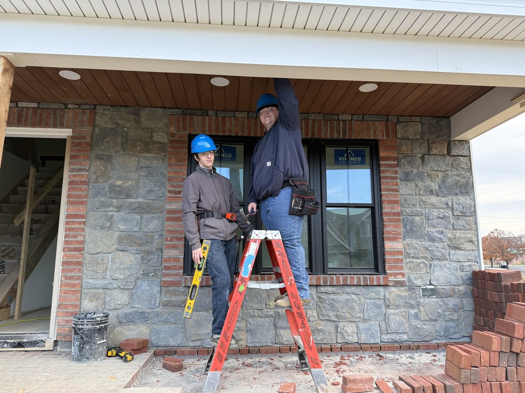 Two Carpentry students stand on a porch area beneath the soffit of the Student Built House. One holds a level while the other stands on a ladder making measurements near the ceiling. Stone and brick exterior finishes surround them.