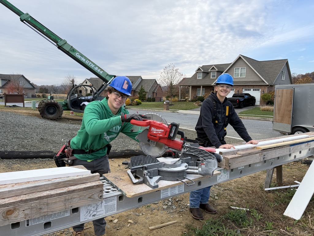 Two Carpentry students wearing blue hard hats and safety glasses stand outside at a job site, smiling while operating a red miter saw set up on a workbench. Houses and construction equipment are visible in the background.