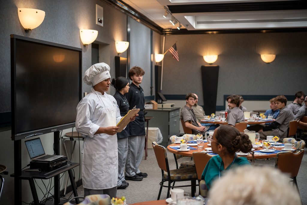 A Culinary Arts student in a white chef uniform stands at the front of a dining room holding a clipboard and speaking to seated students while two hospitality students stand behind them.