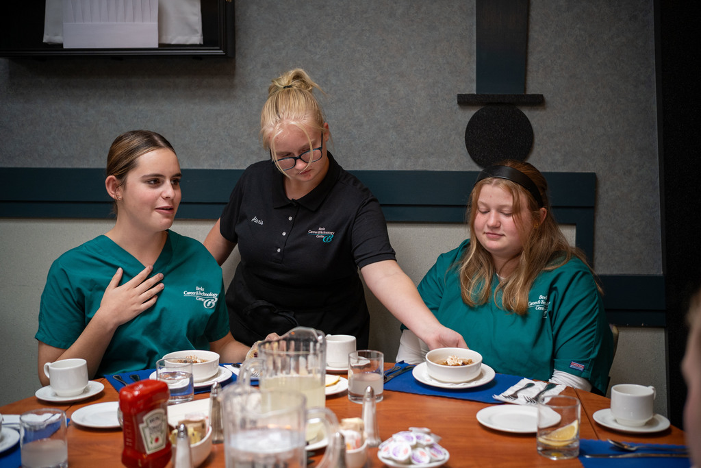 A Hospitality student in a black uniform serves bowls of food to two seated students wearing teal program shirts at a dining table.