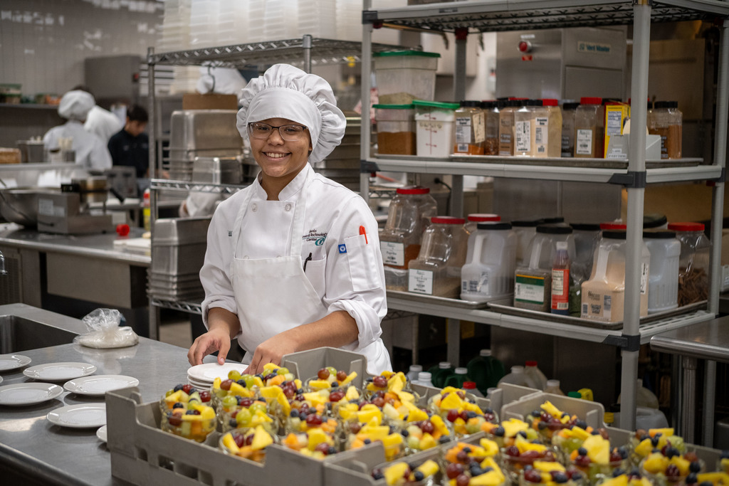 A Culinary Arts student in a white chef uniform smiles while preparing plates beside a cart filled with cups of fresh fruit in a commercial kitchen.