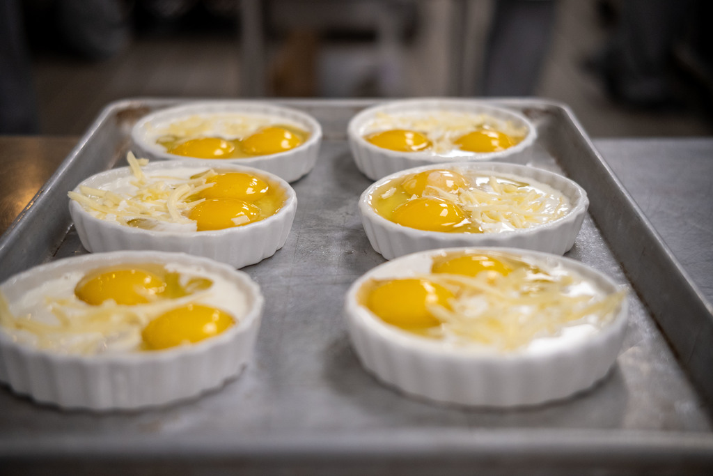 Several white ramekins filled with raw eggs and shredded cheese are arranged on a metal tray, ready to be baked.