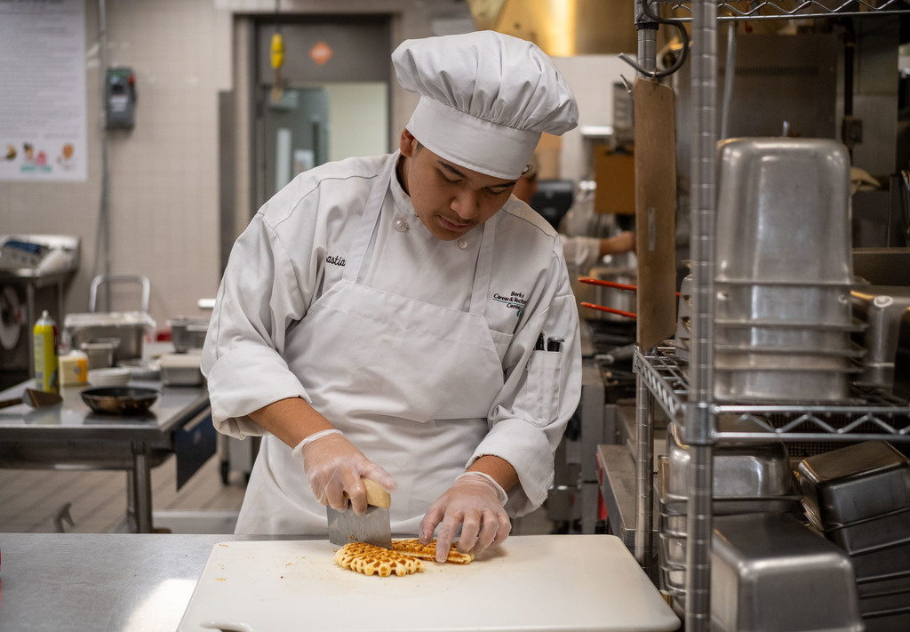 A Culinary Arts student in a white chef coat and hat slices a waffle on a cutting board in a busy commercial kitchen.