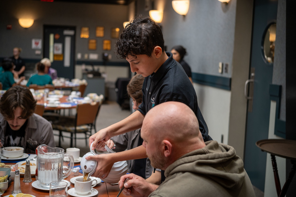 A Hospitality student in a black uniform pours coffee for a guest seated at a breakfast table during the dining experience.