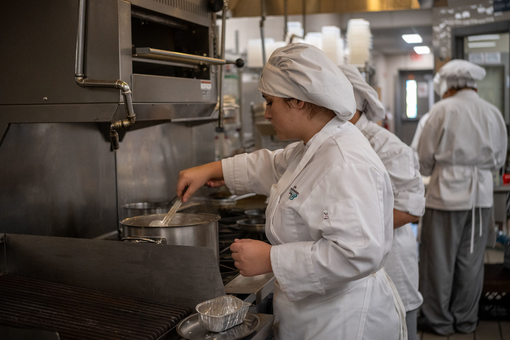 A Culinary Arts student wearing a white chef coat and hat stirs a large pot on the stove in a commercial kitchen, with other students working in the background.