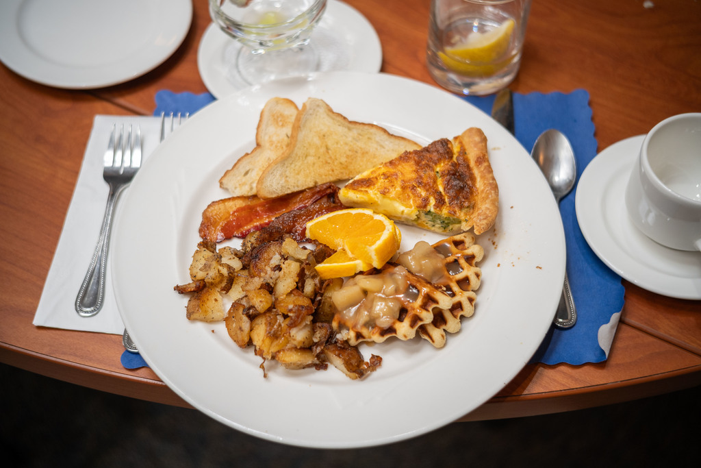 A plated breakfast featuring toast, bacon, home-fried potatoes, a slice of quiche, waffle pieces topped with warm apples, and orange slices sits on a wooden table with silverware and a glass of water.