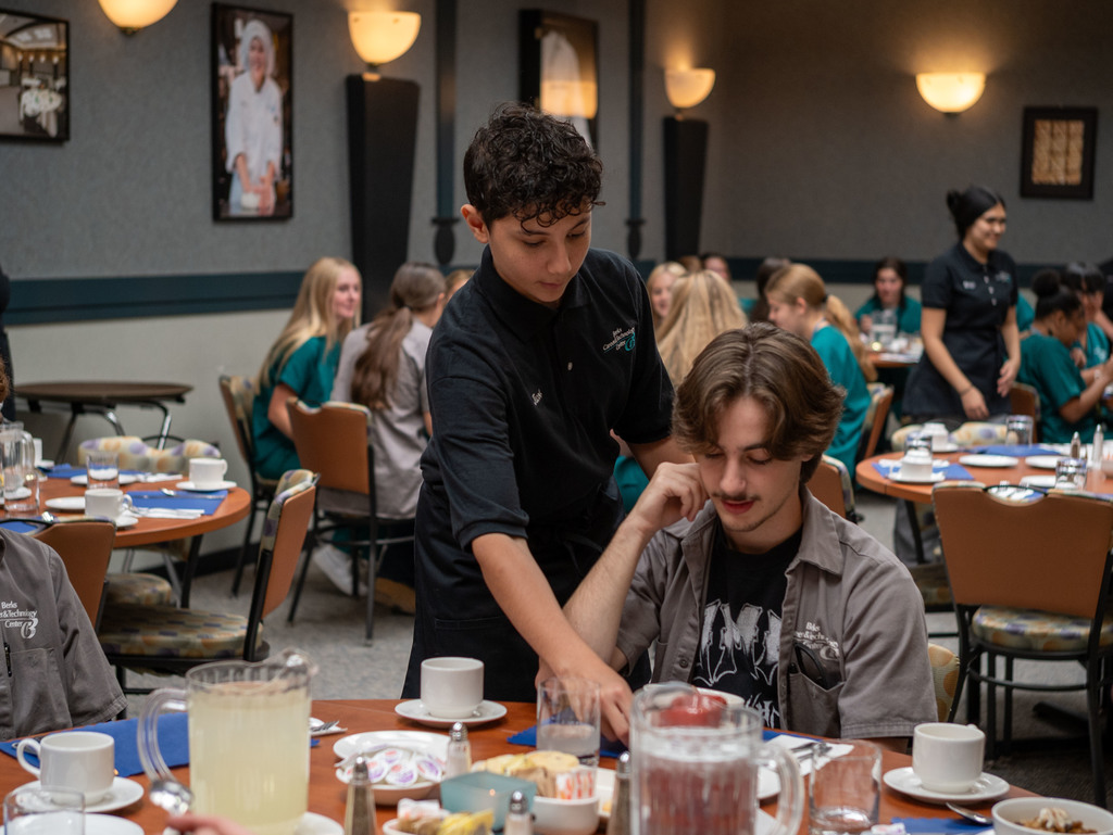 A Hospitality student in a black uniform places a small dish in front of a seated student while others dine in the background.