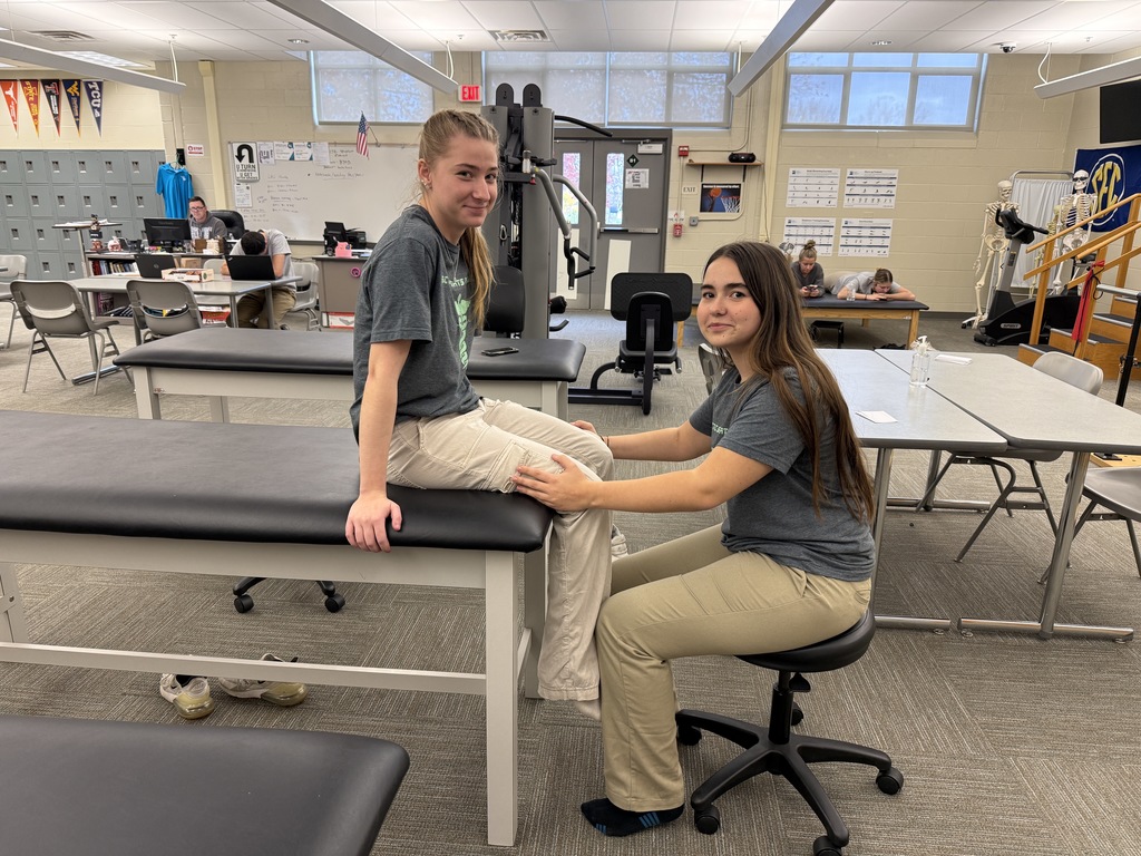 Two Sports Medicine & Rehabilitative Therapy students conduct a knee assessment at a therapy table. One student sits upright with her legs over the edge while the other student applies resistance to her lower leg.