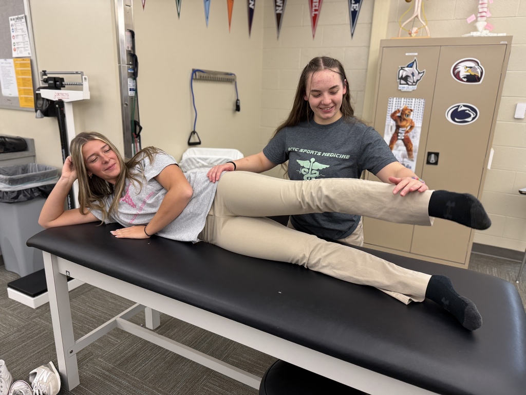 A Sports Medicine & Rehabilitative Therapy student performs a manual muscle strength test on a classmate who is lying on her side on a black therapy table. The student applies gentle resistance to the raised leg while smiling.