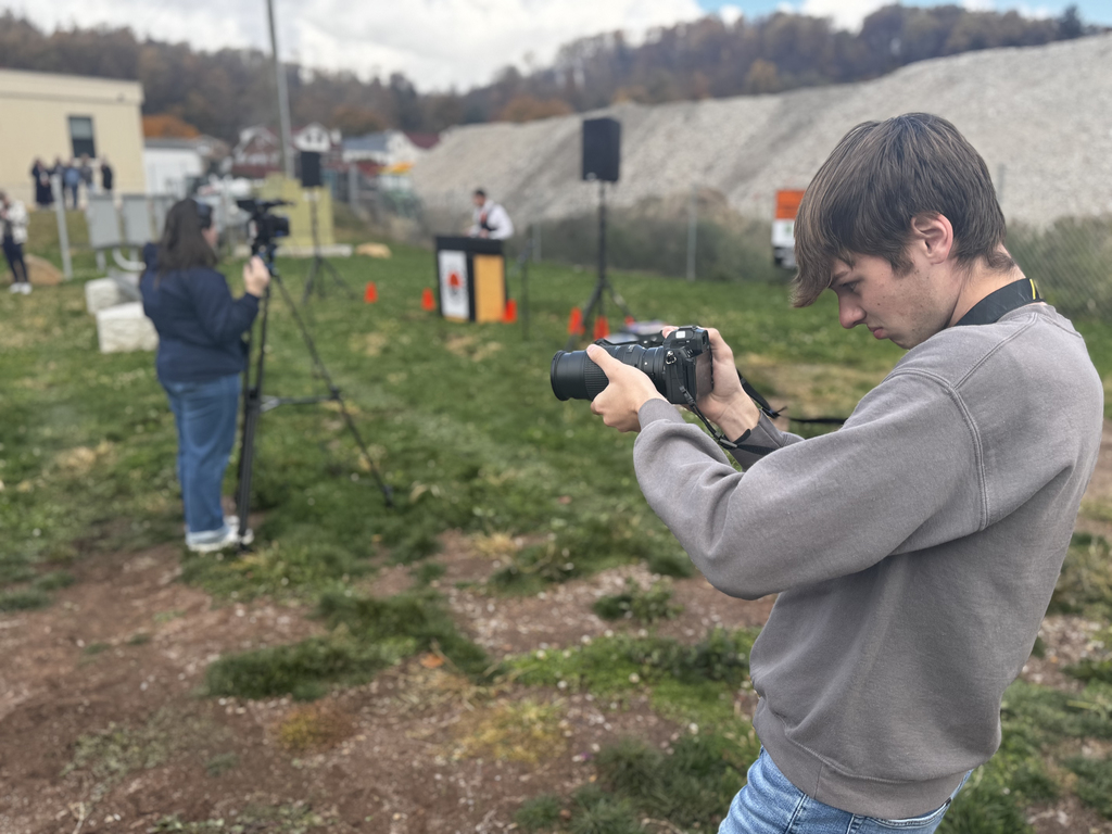 A BCTC student focuses intently while adjusting settings on a Nikon camera, standing on a grassy field during the steel beam signing event. In the background, another student films with a large video camera on a tripod near the podium.
