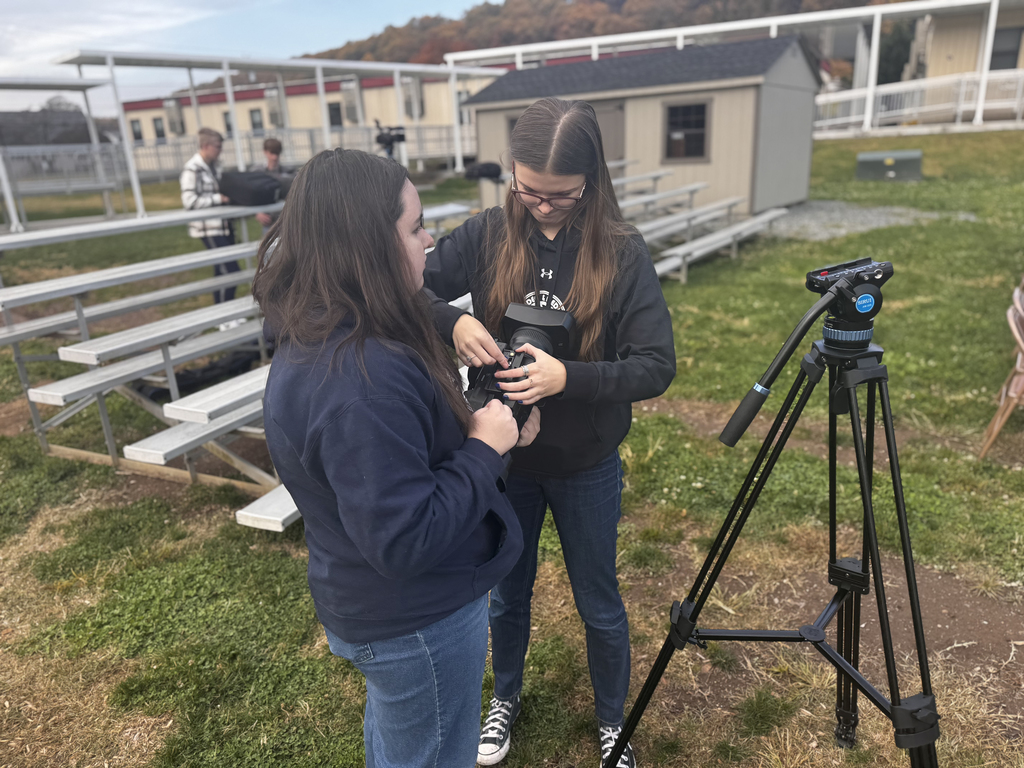 Two female BCTC students stand next to metal bleachers while working together to adjust a camera. One holds the camera body while the other makes adjustments. A tripod with a fluid head is set up beside them, and two male students are visible working with equipment in the background.