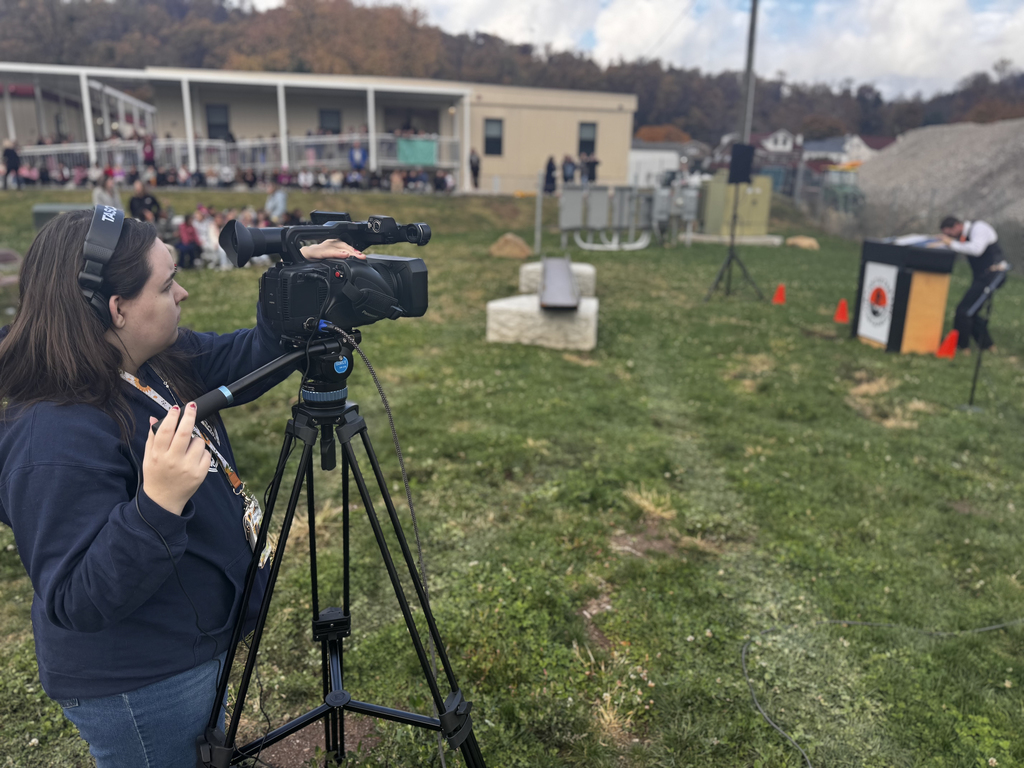 A BCTC student wearing headphones films the steel beam signing event using a professional video camera mounted on a tripod. In the distance, a speaker stands at a podium, and a crowd of students is gathered near the school building behind them.