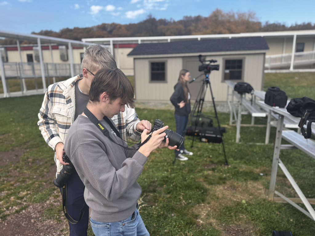 Two BCTC Video & Media Content Production students stand outdoors adjusting settings on a Nikon camera. One student holds the camera while the other leans in closely to assist. In the background, another student stands beside a video camera on a tripod near metal bleachers and a small shed.