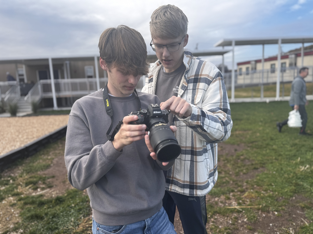 Two BCTC students closely review the screen of a Nikon camera while standing on a grassy area. The student in front holds the camera, and the student behind points toward the controls. School buildings and a person walking in the distance appear in the background.