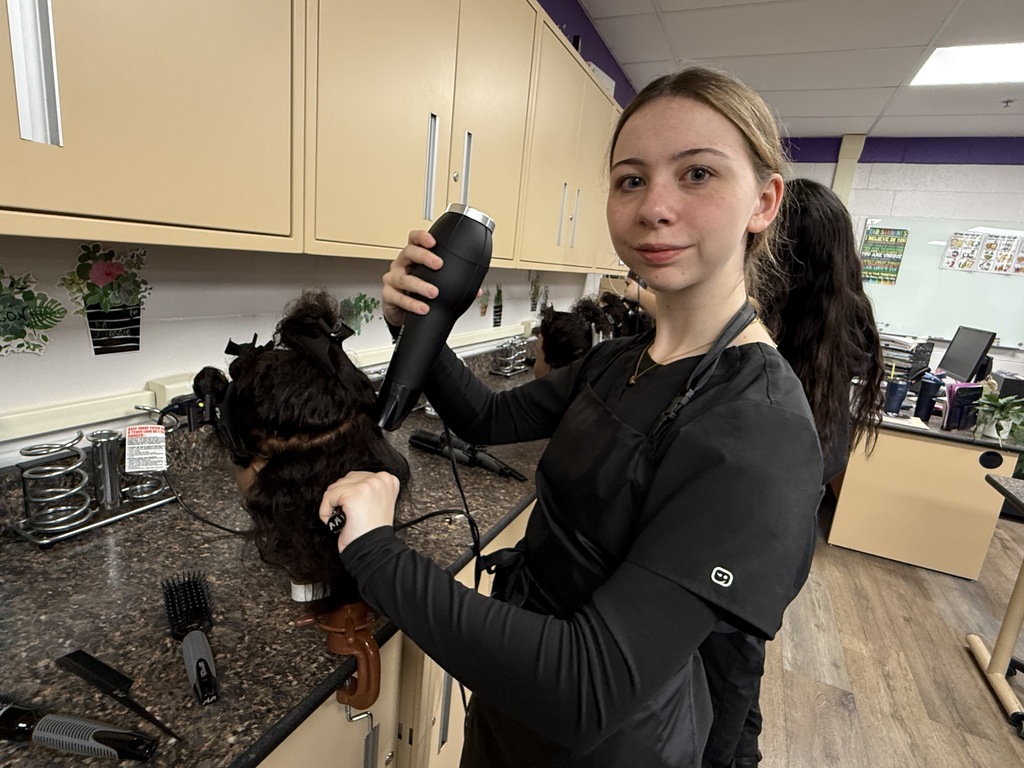 A Cosmetology student with her hair pulled back uses a blow dryer on a mannequin head with textured hair. She looks toward the camera while working at a countertop filled with brushes and tools, with other students working in the background.