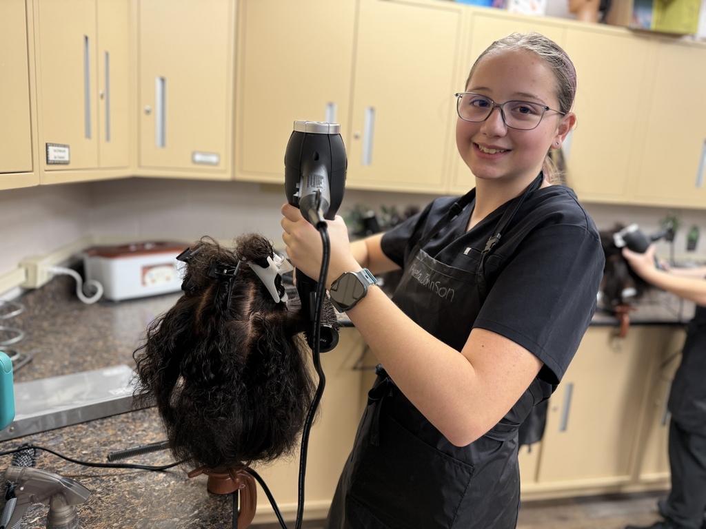 A Cosmetology student wearing black scrubs and glasses uses a blow dryer on a mannequin head with textured hair. She is smiling at the camera while working at a salon station with beige cabinets in the background.