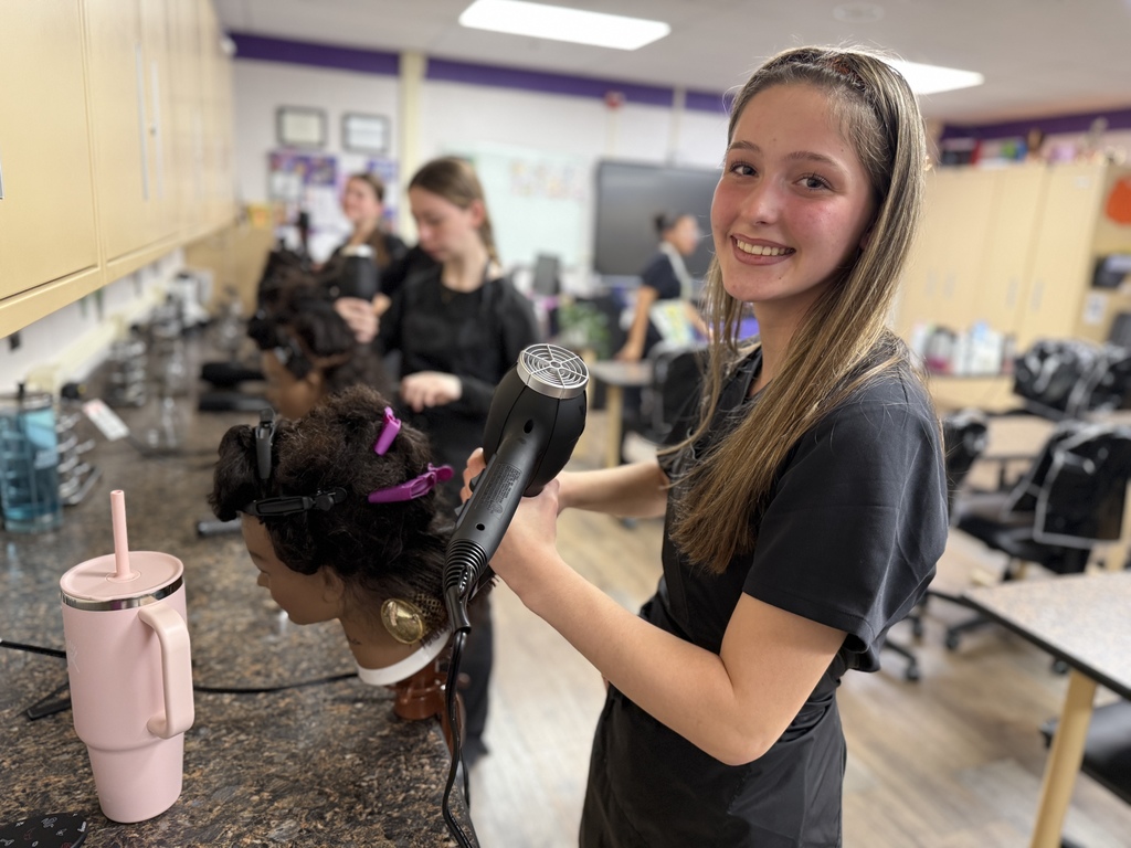 A Cosmetology student with long light brown hair smiles at the camera while holding a blow dryer to a mannequin head with textured hair. Other students work at stations behind her in the salon classroom.