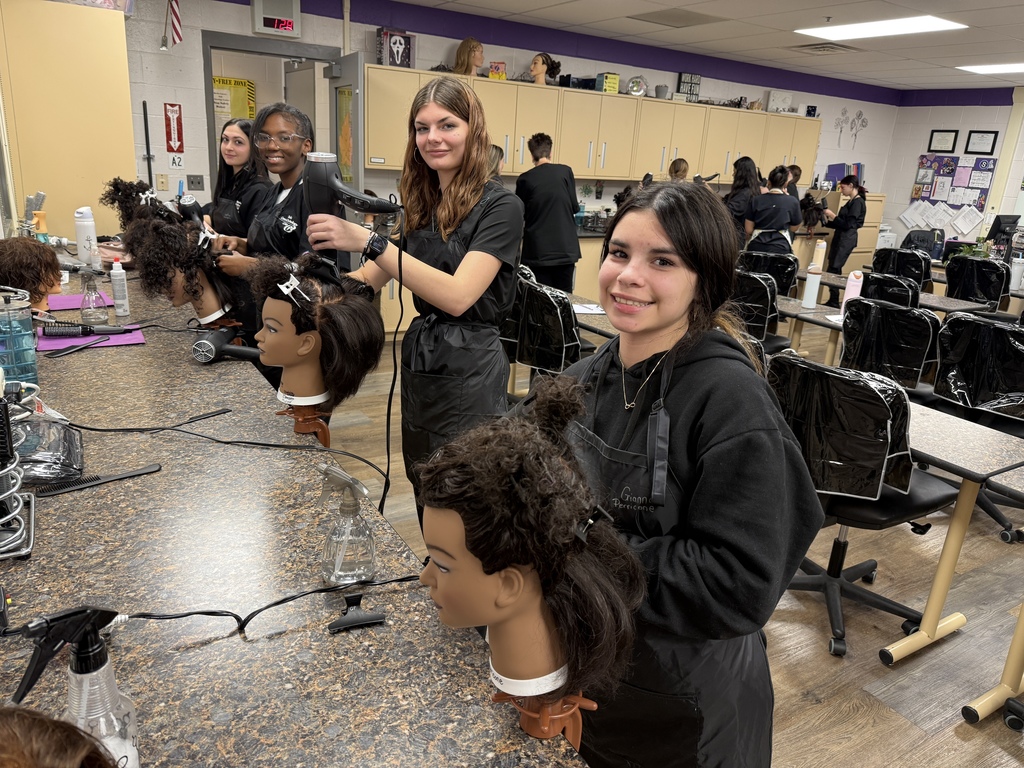 A group of Cosmetology students stand at a long classroom counter, each working on a mannequin head with textured hair. One student in the foreground smiles at the camera while holding a blow dryer, and others work in the background across the salon classroom.