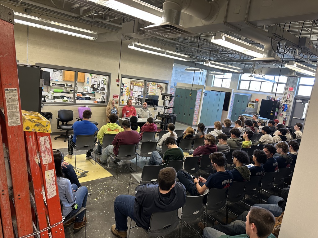 A large group of Construction Cluster students sit in rows of gray chairs inside a workshop classroom, listening to three guest speakers seated at a table in the front of the room. The speakers—a woman and two men—are positioned near a windowed wall that looks into another lab space. The workshop has high ceilings with exposed ducts, fluorescent lighting, and various tools and equipment visible, including a tall red ladder in the foreground on the left.