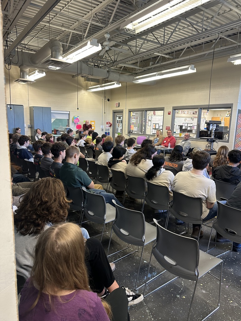 Dozens of Construction Cluster students sit facing three guest speakers at the front of a workshop classroom. The speakers are seated behind a table along a wall of windows that look into another classroom. The room has high ceilings with exposed beams and ducts, overhead fluorescent lighting, and splattered paint marks on the floor. Students fill several rows of gray chairs, listening to the presentation.