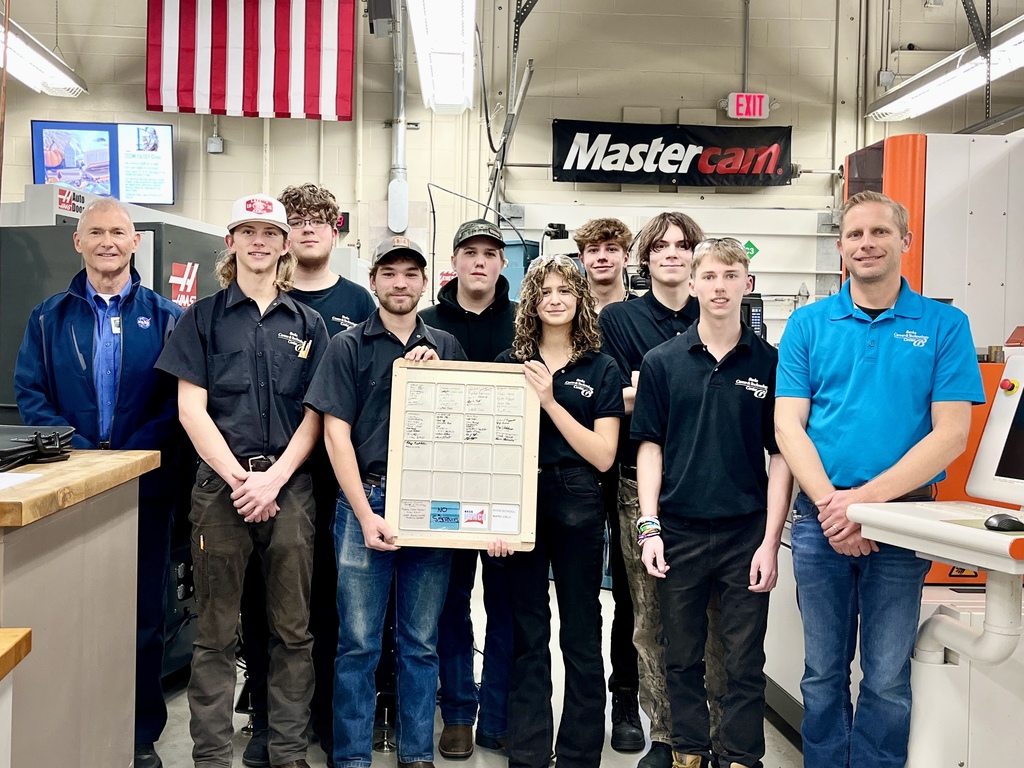 A group of Computerized Machining Technology students stands together in the machining lab posing with their instructor and a NASA representative. One student holds a project planning board in the center, and machining equipment and an American flag are visible behind them.