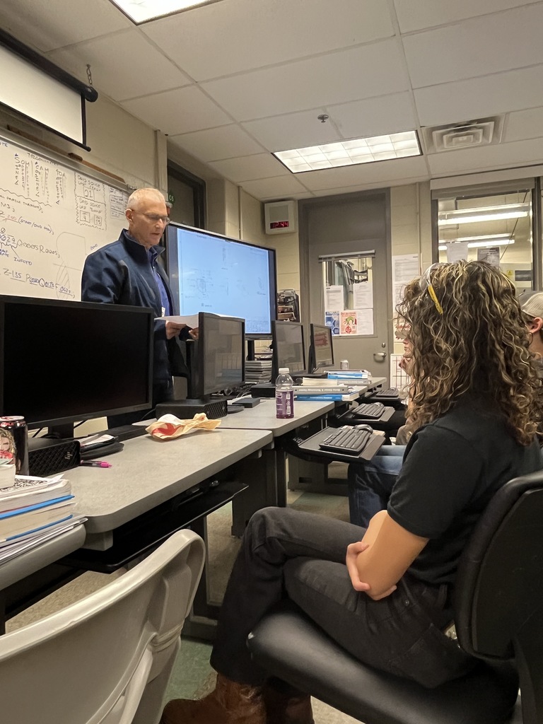 A NASA representative stands near a large screen displaying engineering drawings while students sit at computers listening. A student in the foreground, wearing dark clothing and curly hair, is seated and facing forward.