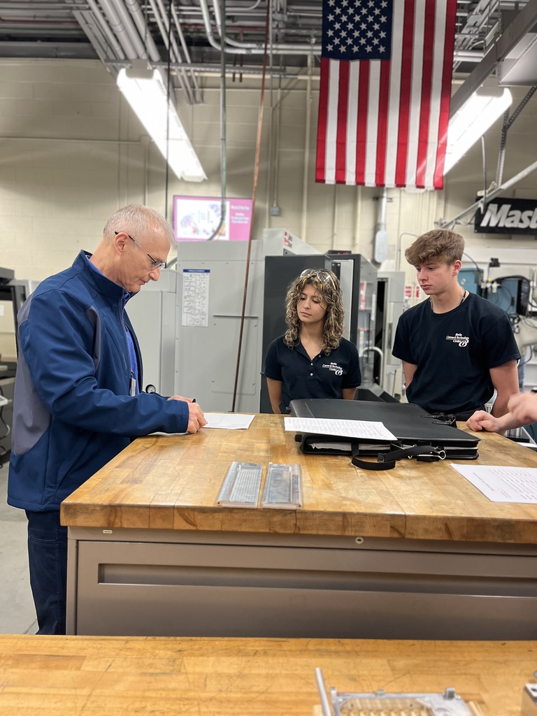 A NASA representative reviews papers with two Computerized Machining Technology students at a wooden workbench in the machining lab. The students stand across from him while CNC equipment and an American flag hang in the background.