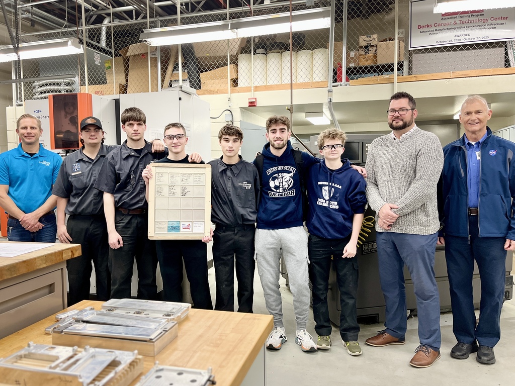 A group of Computerized Machining Technology students stands together in a machining lab, posed in a line between two instructors and a NASA representative. One student in the center holds a large project planning board. CNC machines and shop equipment are visible in the background.