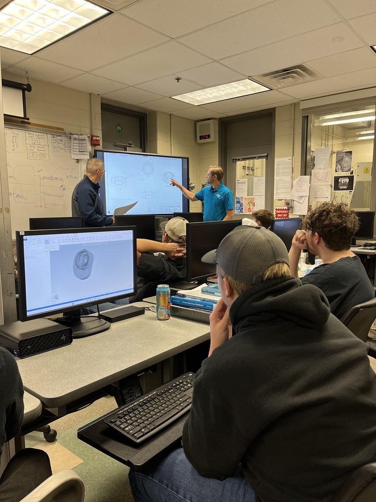 A NASA representative and a BCTC instructor stand at the front of a computer lab reviewing a technical drawing displayed on a large screen. Students seated at computer stations look on as a 3D model appears on their monitors.