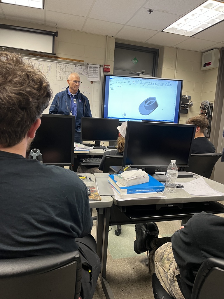 A NASA representative stands in front of a projected 3D CAD model as students sit at computer stations watching. Technical notes and sketches cover the whiteboard behind him.