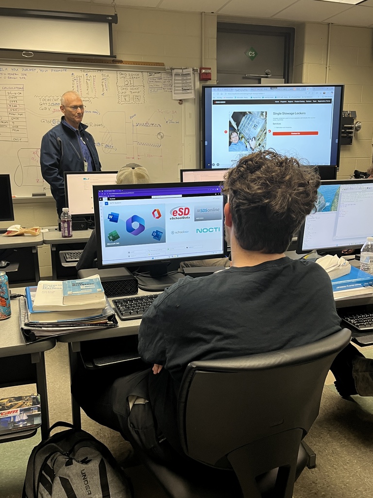 A NASA representative stands at the front of a classroom, speaking to students seated at desktop computers. A large monitor displays an image of space equipment, and handwritten machining notes cover the whiteboard behind him.