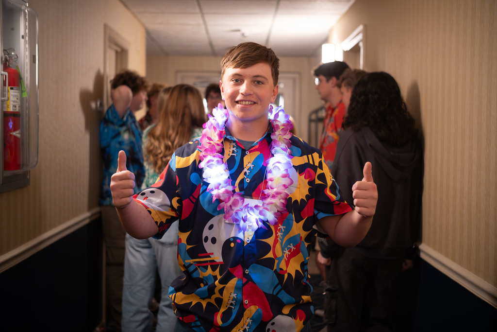 A student wearing a colorful Hawaiian shirt and a glowing LED lei stands in a hotel hallway giving two thumbs up. Other students in bright shirts are gathered and talking behind him.