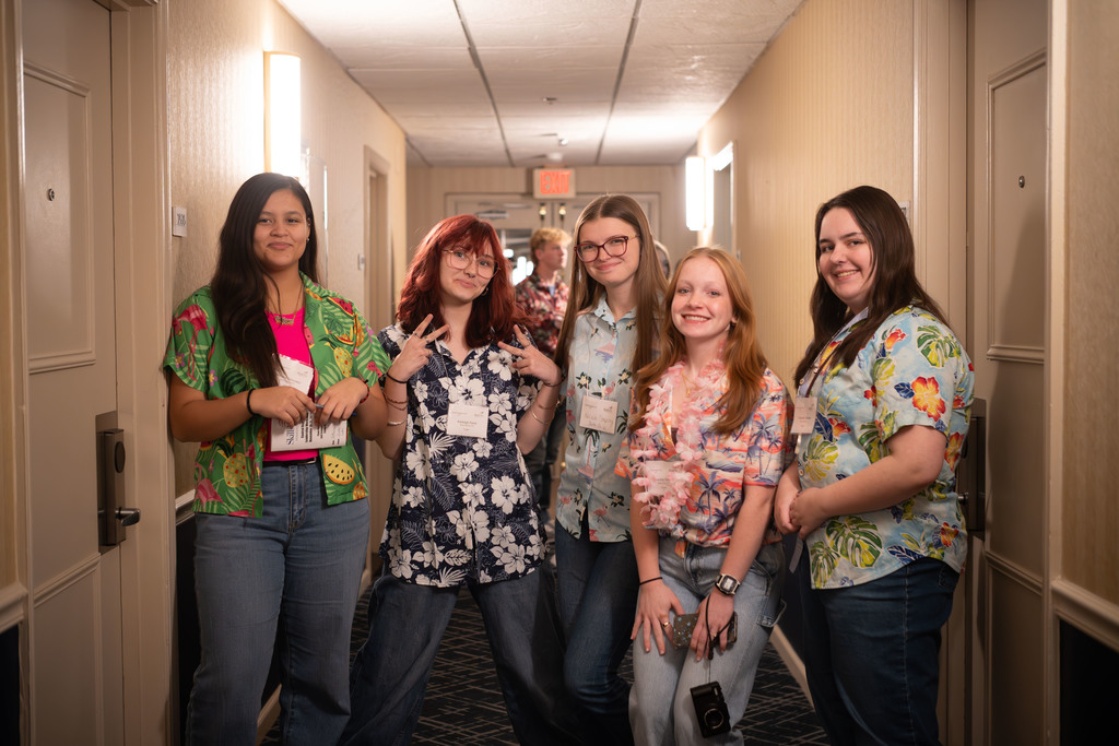 A group of five students stand in a hotel hallway wearing bright Hawaiian shirts. They smile at the camera, and one student in the middle holds up a playful peace sign. The hallway is softly lit with beige walls and room doors on both sides.