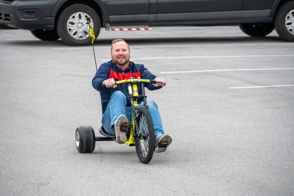 An adult rides a low-to-the-ground drift tricycle in a parking lot, smiling as he pedals. Parked vehicles are visible in the background.