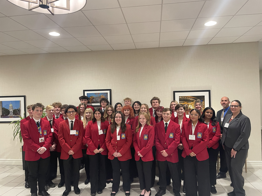 A large group of SkillsUSA students and instructors pose together in a hotel lobby. They are dressed in red SkillsUSA blazers and black pants, arranged in several rows with framed artwork and soft lighting behind them.