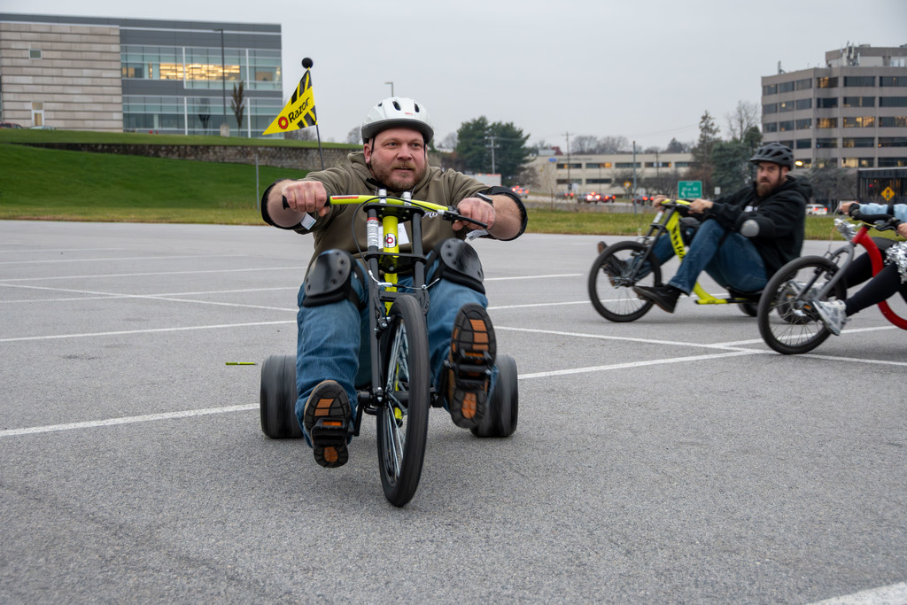 An adult wearing a helmet and knee pads rides a drift tricycle in a parking lot. Two other riders follow behind him, and buildings line the background.