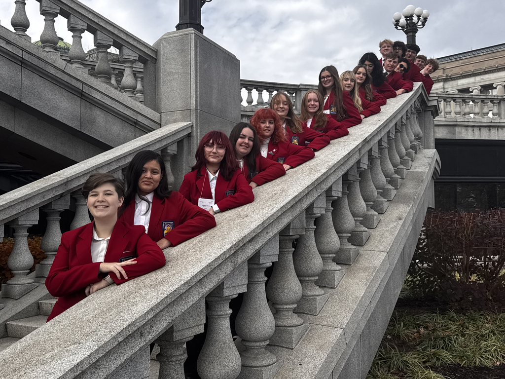 A line of SkillsUSA students in red blazers stand along a stone banister outside the Pennsylvania State Capitol. They are positioned diagonally down the railing, smiling toward the camera.