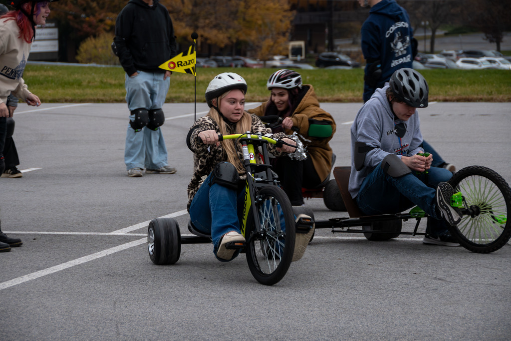 A student wearing a helmet and a leopard-print jacket rides a drift tricycle during an outdoor activity. Other students on tricycles and standing nearby are visible behind her.