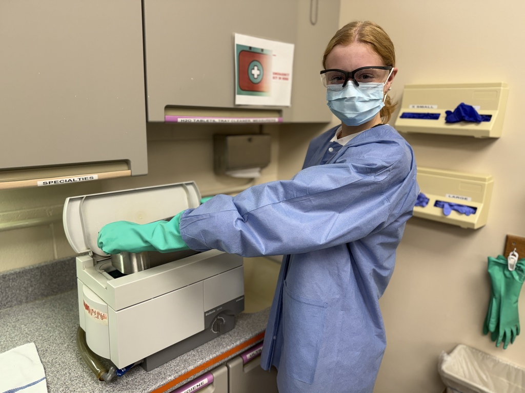 A Dental Occupations student wearing safety glasses, a face mask, a blue protective gown, and green gloves loads instruments into an ultrasonic cleaner in a clinical lab setting. Cabinets, labeled storage areas, and wall-mounted glove dispensers are visible in the background.