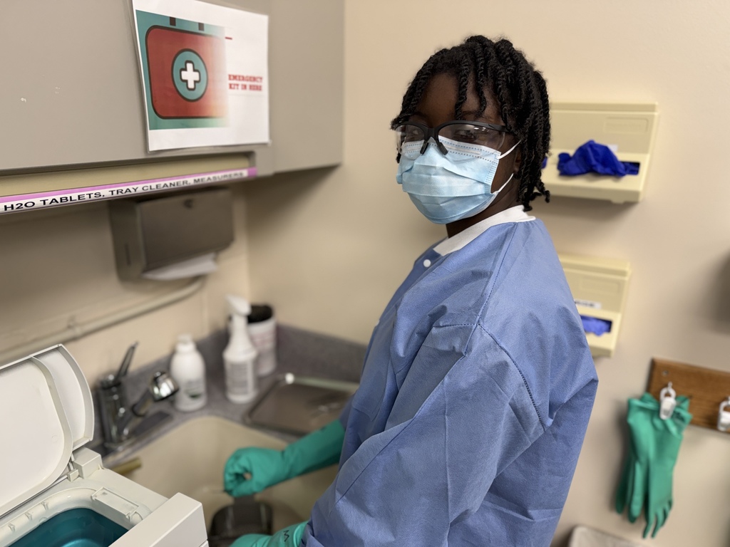 A Dental Occupations student wearing safety glasses, a face mask, a blue protective gown, and green gloves stands at a workstation next to an ultrasonic cleaner. The student looks toward the camera while working with dental instruments. Cabinets, cleaning supplies, and glove dispensers are visible behind them.