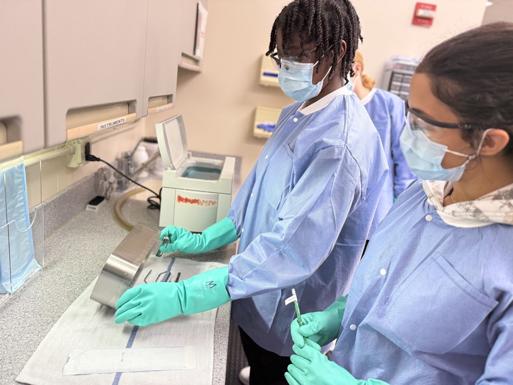 Two Dental Occupations students wearing protective gowns, masks, eyewear, and green gloves stand at a counter preparing dental instruments. One student holds a metal tray while the other holds a dental tool. An ultrasonic cleaner and labeled cabinets are visible on the counter.
