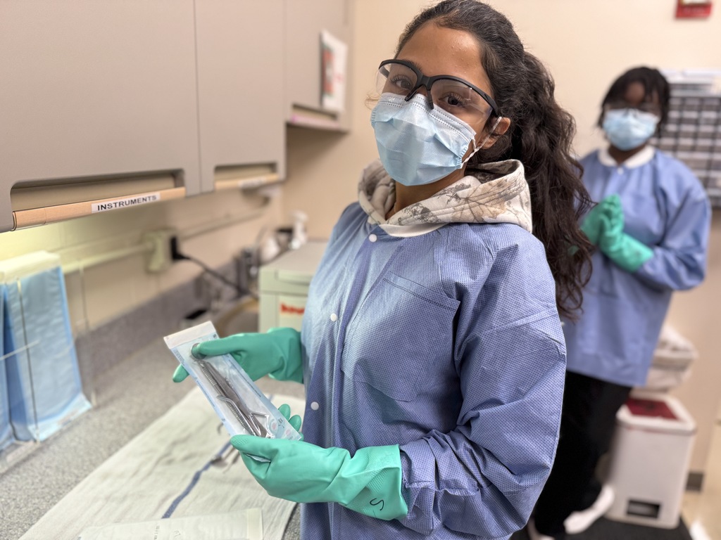 A Dental Occupations student wearing safety glasses, a face mask, a blue protective gown, and green gloves holds a sealed sterilization pouch containing dental instruments. Another student stands behind them near wall-mounted glove dispensers. The setting is a clinical lab workspace.