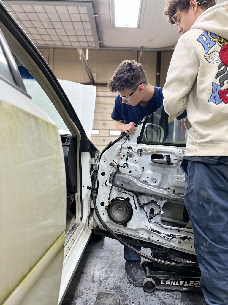 Two Automotive Collision Repair Technology students work on removing the interior panel of a car door. One student leans in closely while inspecting wiring, and the other stands beside him. Both are wearing safety glasses inside an automotive shop.