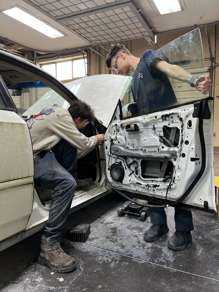 Two Automotive Collision Repair Technology students work together to disassemble a car door. One student is seated and working with tools near the lower section of the door, while the other holds the glass window upright. The scene takes place inside an automotive shop.