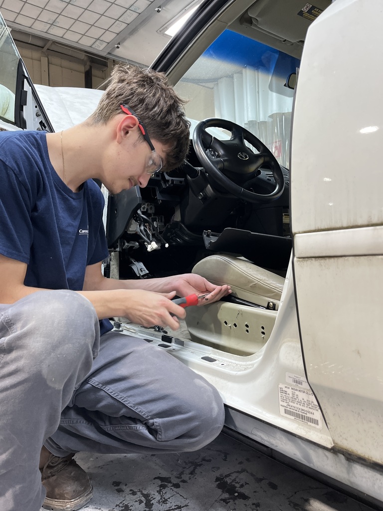 An Automotive Collision Repair Technology student wearing safety glasses kneels beside an open vehicle and uses a screwdriver to remove a component under the front seat. The car’s interior is partially disassembled.