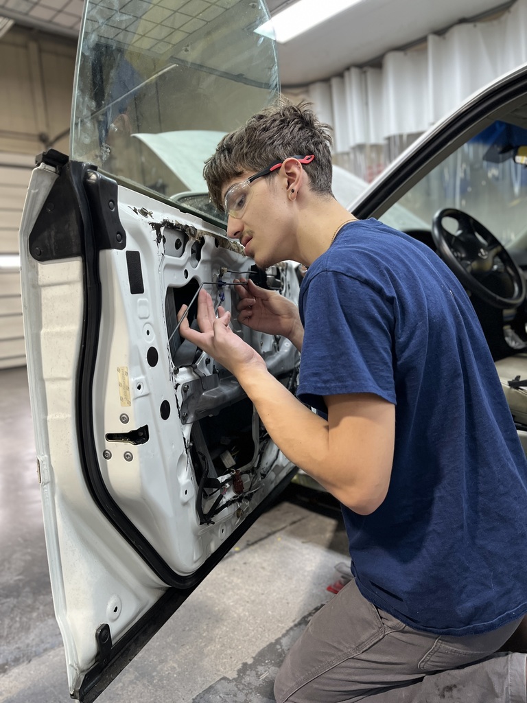 An Automotive Collision Repair Technology student wearing safety glasses kneels beside an open car door and uses a tool to disconnect wiring inside the door frame. The interior panel has been removed to expose mechanical components.