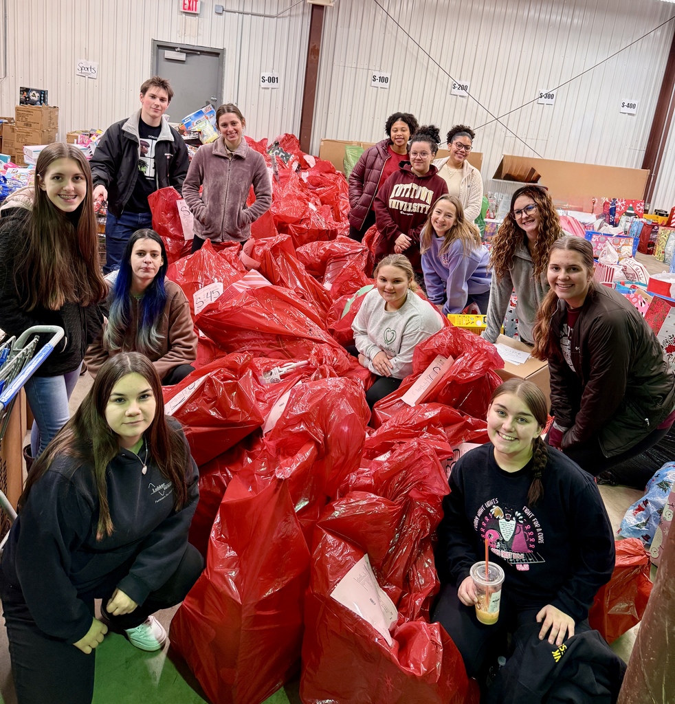 A large group of students poses in a warehouse sitting and kneeling beside stacks of oversized red donation bags. They are smiling and surrounded by rows of sorted gift bags marked with category labels on the walls.