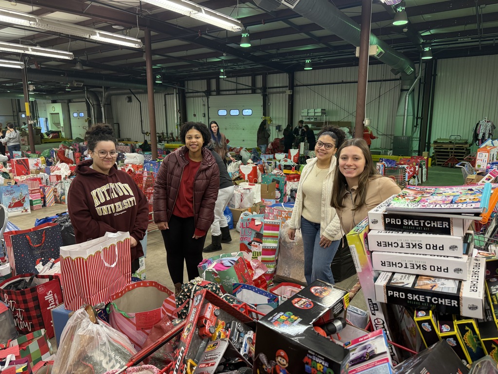 Four students stand inside a warehouse among stacks of wrapped toys and bags. They are smiling while surrounded by colorful holiday donations including boxes, bags, and gift sets.