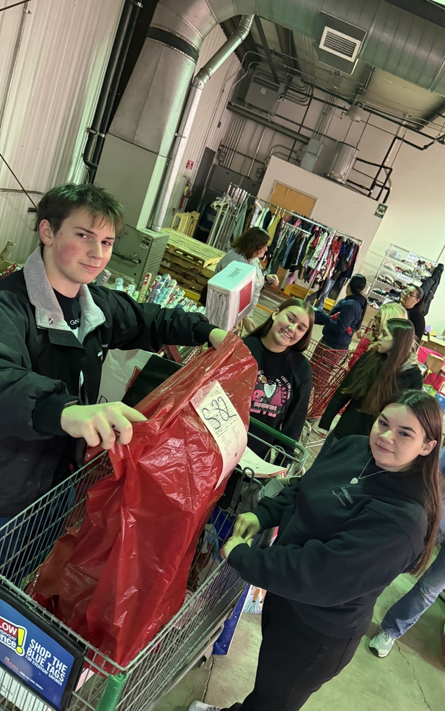 Three students stand beside a shopping cart inside a warehouse, placing a large red bag inside it. Shelves, clothing racks, and other volunteers are visible in the background as they help sort donations.