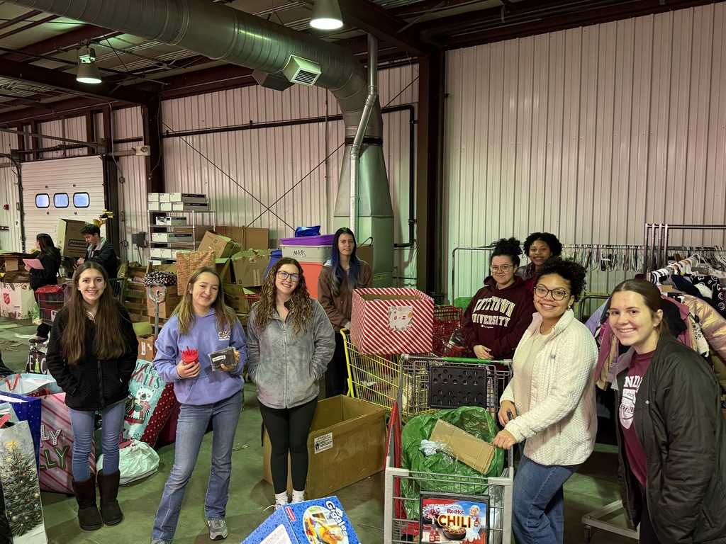 A group of students stands inside a warehouse surrounded by holiday donation items. They are smiling and pushing donation carts filled with wrapped gifts and winter clothing, with rows of items visible behind them.